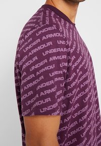 Lila t-shirt med kort ärm som har upprepande texten "UNDER ARMOUR" i ljusare lila. Mjuk tyg, rundhalsad design och standard passform.