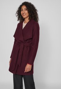 Burgundijas krāsa mētelis ar platu atlokiem, jostu viduklī un sānu kabatām. Izgatavots no mīksta auduma. Apvienots ar vieglu džemperi un melniem biksēm.