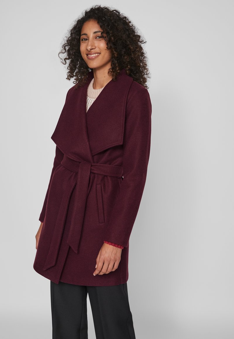 Burgundijas krāsa mētelis ar platu atlokiem, jostu viduklī un sānu kabatām. Izgatavots no mīksta auduma. Apvienots ar vieglu džemperi un melniem biksēm.