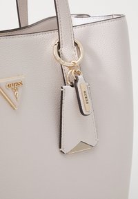 Sac en cuir taupe avec des accents de quincaillerie dorée, surface texturée et une étiquette signature. Comprend un détail de logo triangulaire.