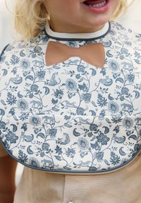 Bibben har en blommönstrad design med blå och grå nyanser, en ljus bakgrund och en brun vinylrosett som accent vid halsringningen.