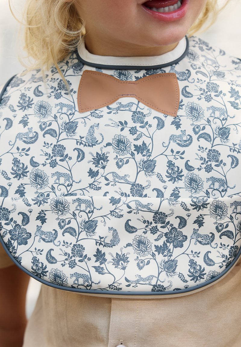 Bibben har en blommönstrad design med blå och grå nyanser, en ljus bakgrund och en brun vinylrosett som accent vid halsringningen.