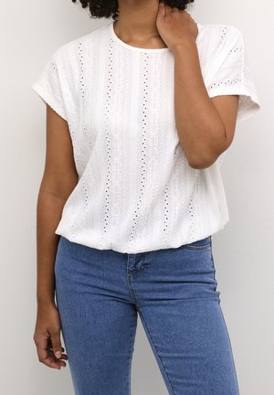 Kobieta w białej bluzce z haftem angielskim z krótkim rękawem i niebieskich dżinsach, jedna ręka dotyka szyi na jednolitym tle.