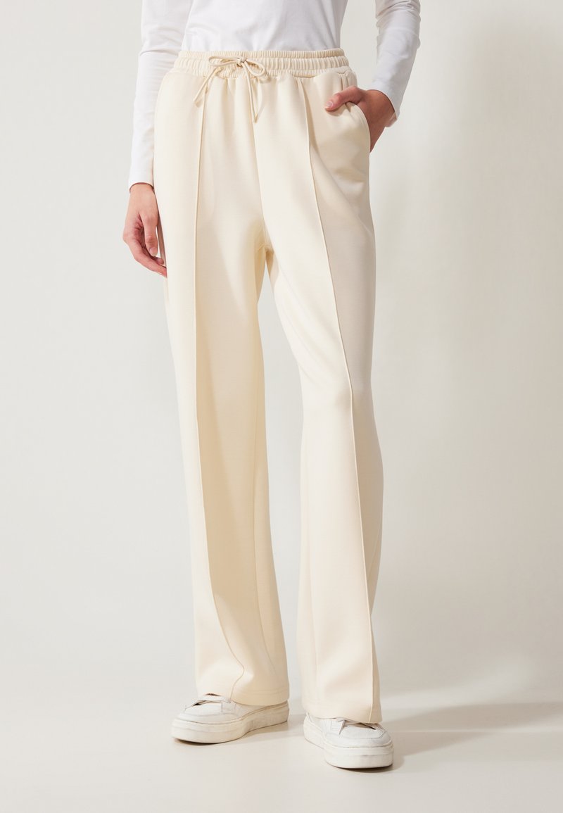 Pantalons larges de couleur crème avec une taille ajustée, des poches latérales et une texture de tissu lisse. Associés à des baskets blanches.