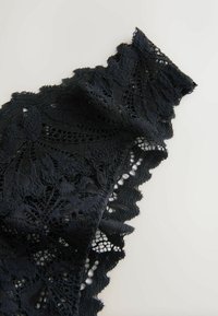 Mustekaskanka, joka esittää kukkakuvioita, aaltoilevia reunoja ja tekstuurista pintaa, on aseteltu vaalealle taustalle.