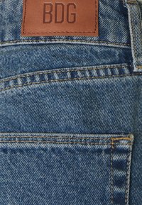 Jeans i denim i blått med en ljus tvätt, med en brun läderpatch som har texten "BDG" och kontrasterande gul sömnad på bakfickan.