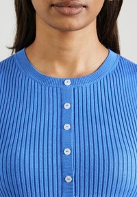 Vrouw in een lichtblauwe, geribbelde knooptrui, met een open hals, lippen met matte, bruine lippenstift en donker haar tegen een neutrale achtergrond.