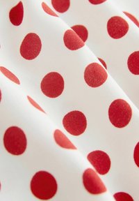 Biely látka s vzorom rovnomerne rozmiestnených červených bodiek, zobrazená v detailnom zábere s jemnými záhybmi.