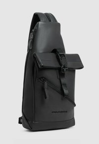 Zwarte schoudertas gemaakt van glad synthetisch materiaal, met een klep met gesp, een ritsvak en een schuine vormgeving. Minimalistische merkuitstraling zichtbaar.