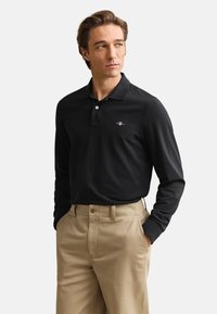 Hombre con camiseta polo negra de manga larga y pantalones beige, de pie con las manos en los bolsillos, mirando ligeramente hacia un lado.