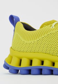 Κίτρινο αθλητικό παπούτσι με υφασμάτινη επάνω επιφάνεια και μπλε λεπτομέρειες στη φτέρνα. Διαθέτει χοντρή κίτρινη σόλα με κυκλικές εγκοπές για υποστήριξη.