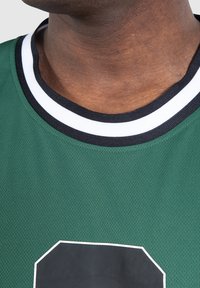 Detailní záběr na krk a horní část hrudníku osoby, která nosí zelený sportovní dres s černým a bílým límečkem a velkým černým číslem orámovaným bílou barvou.