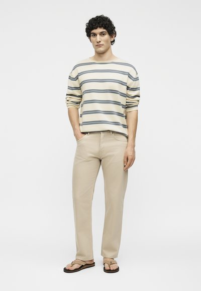 Homme aux cheveux bouclés portant un pull beige rayé, un pantalon beige et des sandales beige, debout avec une main dans la poche.
