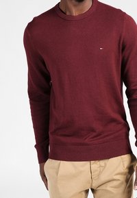 Burgundy stickad tröja med rundad hals, ribbade ärmslut och nederkant. Har en liten logotyp i blått och vitt på bröstet.