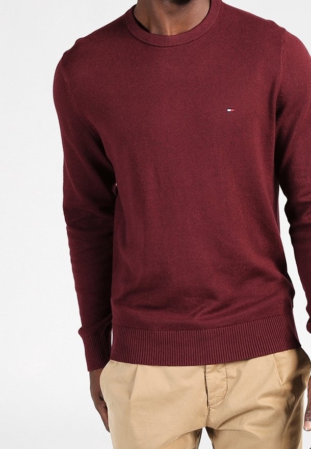 Burgundy stickad tröja med rundad hals, ribbade ärmslut och nederkant. Har en liten logotyp i blått och vitt på bröstet.