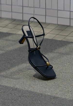 Svart remsandal med høye hæler, firkantet tå, gullspenne og gullperledetalj på tåremsen, plassert på asfalt nær flislagt vegg.