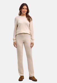 Beżowy sweter dziany i dopasowane spodnie, oba o miękkiej fakturze i z prążkowanymi mankietami, zestawione z brązowym obuwiem dla kontrastu.