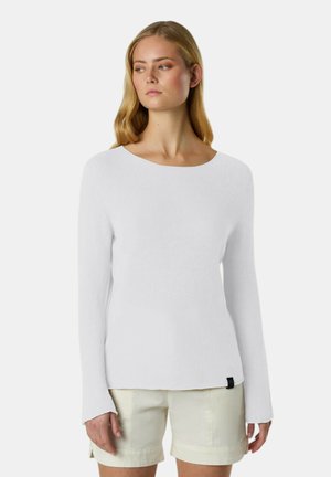 Donna con lunghi capelli biondi, indossa un top bianco a maniche lunghe e pantaloncini beige chiaro, guarda leggermente verso sinistra su uno sfondo semplice.