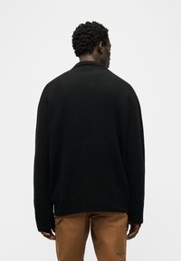 Άντρας με κοντά κατσαρά μαλλιά που κοιτάζει προς τα πίσω, φορώντας μαύρο πουλόβερ και καφέ παντελόνι με πλαϊνές τσέπες, σε λευκό απλό φόντο.