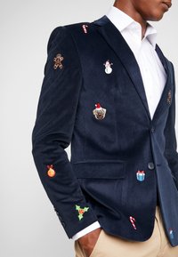 Granatowa marynarka z żakardu z różnorodnymi kolorowymi haftami w tematyce świątecznej, w tym z piernikiem, bałwankiem, psem rasy pug oraz akcentami w formie lasek cukrowych.