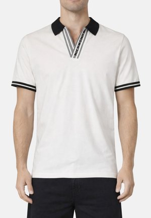 Uomo che indossa una polo bianca a maniche corte con colletto nero, bordatura a righe a V e striscia nera sulle maniche, abbinata a pantaloni neri.