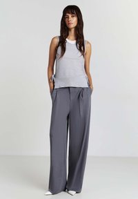 Canotta grigia chiara in knit con lacci laterali, abbinata a pantaloni a gamba larga grigi a vita alta. La modella ha i capelli lunghi e uno sfondo neutro.