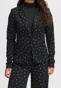 Kvinna som bär en svart dräkt med små vita hjärtmönster, tillsammans med en svart polotröja.