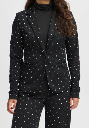 Femme portant un costume noir avec de petits motifs en forme de cœur blancs, associé à un col roulé noir.