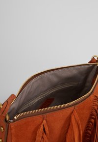 Brązowa torebka z zamszu z zapięciem na zamek, wewnętrzną kieszenią na zamek i frędzlami. Wykończona gładkim, jasnoszarym materiałem.