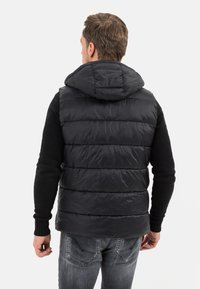 Schwarze Steppweste mit Kapuze, die über gesteppte Paneele, eine glatte Textur und kontrastierende Strickärmel verfügt, kombiniert mit grauen Jeans.