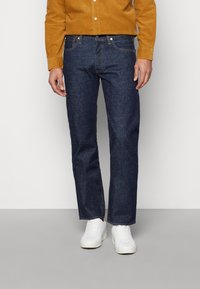 Mörkblå denimjeans med rak passform, åtföljda av en guldkord-uppknäppt skjorta. Vita sneakers fulländar outfiten.
