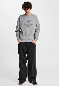 Grijze trui met een ronde hals en manchetten, met een donkerblauw gedrukt logo en tekst. Draagt losse, zwarte jeans en donkere schoenen.