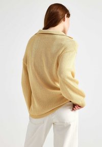 Kvinde med langt rødbrunt hår iført en løs beige striktrøje og off-white bukser, vendt væk med den ene hånd i lommen.