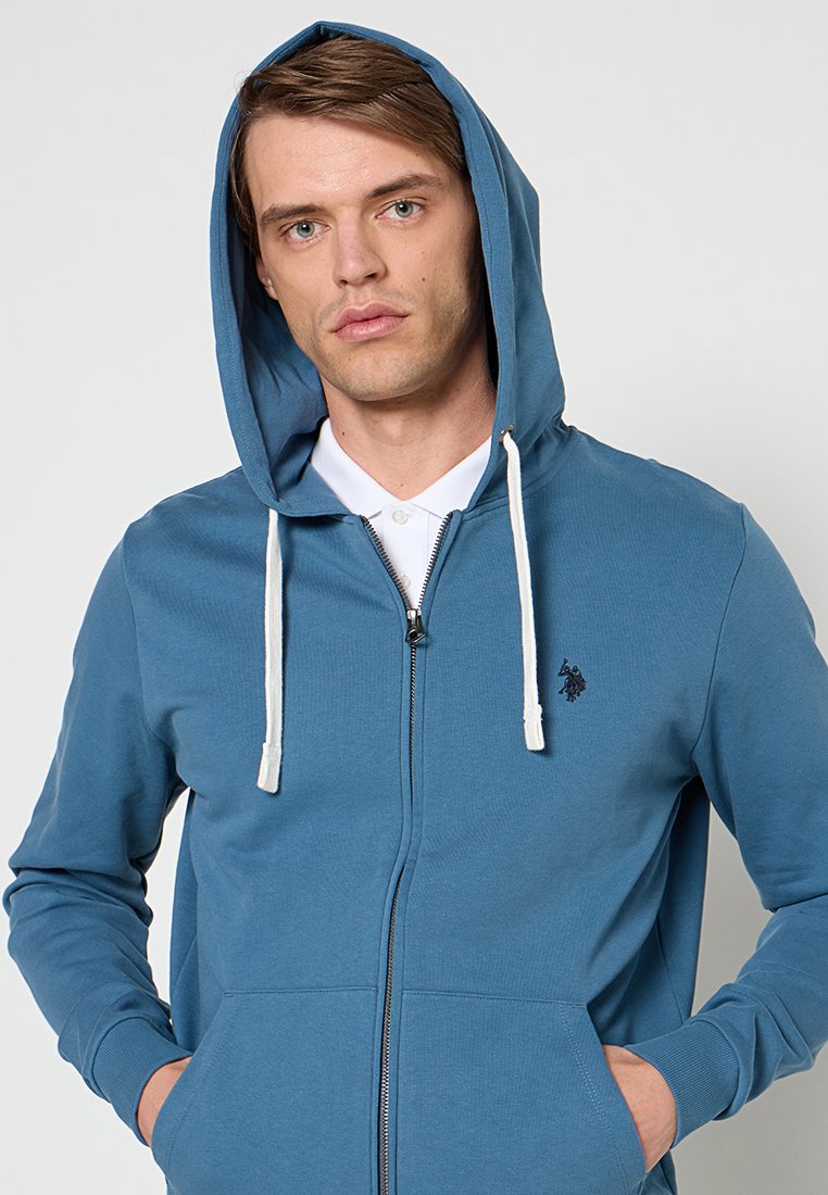 Młody mężczyzna w niebieskiej bluzie z zamkiem błyskawicznym, z białymi sznurowadłami i rękami w kieszeniach, patrzący prosto przed siebie na tle jednolitego tła.