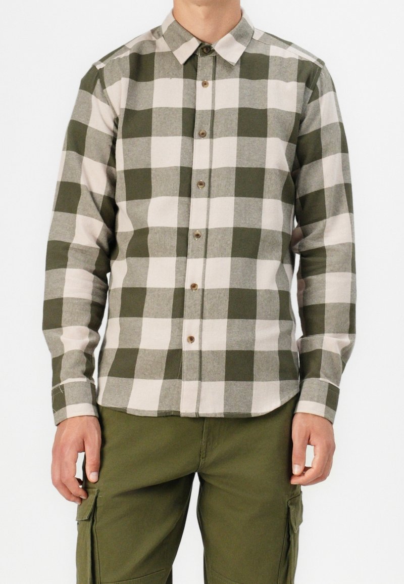 Homme portant une chemise à carreaux verte et blanche à manches longues et un pantalon cargo vert olive, debout avec les bras détendus.