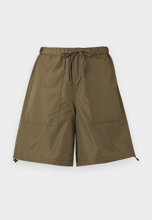 Άνδρας με ελιά πράσινο σορτς με κορδόνι, καφέ μακρυμάνικο πουκάμισο, λευκές κάλτσες και καφέ δερμάτινα παπούτσια-φανταράκια στέκεται μπροστά σε μια απλή φόντο.