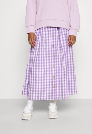 Person trägt ein pinkes Sweatshirt, einen lila-weiß karierten, geknöpften Rock, weiße Socken und weiße Turnschuhe und steht vor einem einfarbigen Hintergrund.