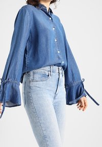 Denim-paita keskisinisessä sävyssä, jossa on rento istuvuus, nappisulku edessä ja levenevät hihat, jotka on sidottu ranteista. Yhdistetty vaaleansinisiin farkkuihin.