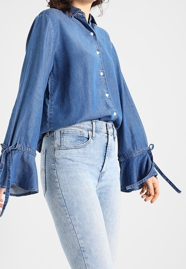 Denim-paita keskisinisessä sävyssä, jossa on rento istuvuus, nappisulku edessä ja levenevät hihat, jotka on sidottu ranteista. Yhdistetty vaaleansinisiin farkkuihin.