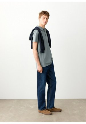 Jonge man staand binnenshuis, gekleed in een grijs poloshirt, donkere spijkerbroek, lichtbruine schoenen en een donkerblauwe trui over zijn schouders gedrapeerd.