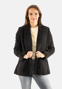 Giovane donna che indossa un blazer in velluto a costine nero sopra una camicia crema con bottoni e jeans blu sbiaditi, in piedi contro uno sfondo semplice.