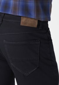 Black denim pants with a textured brown leather patch at the back, featuring a distinctive pocket design and contrasting white stitching.