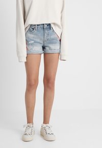 Shorts en denim bleu clair avec des détails déchirés, ourlet effiloché et coupe décontractée, associés à des baskets blanches lacées et un pull beige.
