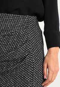 Jupe à motif pied-de-poule noir et blanc avec une finition texturée, dotée d'une taille ajustée et de pliages, avec une main partiellement visible sur le côté.