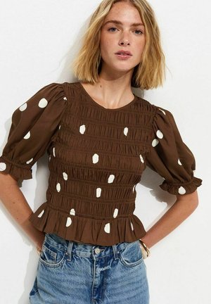 Femme portant un chemisier marron smocké à pois blancs avec un ourlet à volants, associé à un jean en denim bleu taille haute.