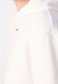 Vue latérale d'une personne portant un sweat à capuche blanc cassé avec cordons et texte noir partiellement visible près de la poitrine.