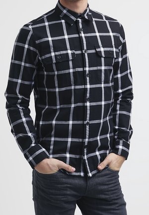Homme portant une chemise à manches longues à carreaux noirs et blancs avec des poches poitrine à l’avant, mains dans les poches d’un pantalon foncé.