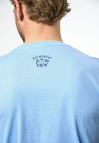 Svetlo modra bombažna majica s kratkimi rokavi, ki na hrbtni strani prikazuje okroglo temno modro grafiko z napisom "AUTENTIČNO 1890 ZDA".