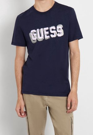 Mężczyzna ubrany w granatowy t-shirt z pogrubionym napisem "GUESS" i beżowe spodnie typu cargo, stojący na tle jednolitego jasnego tła.
