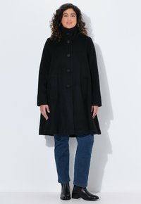 Abrigo negro de mitad de longitud con cuello alto, cierre de botones, bolsillos laterales y una silueta holgada, combinado con vaqueros azules y botas negras de tobillo.